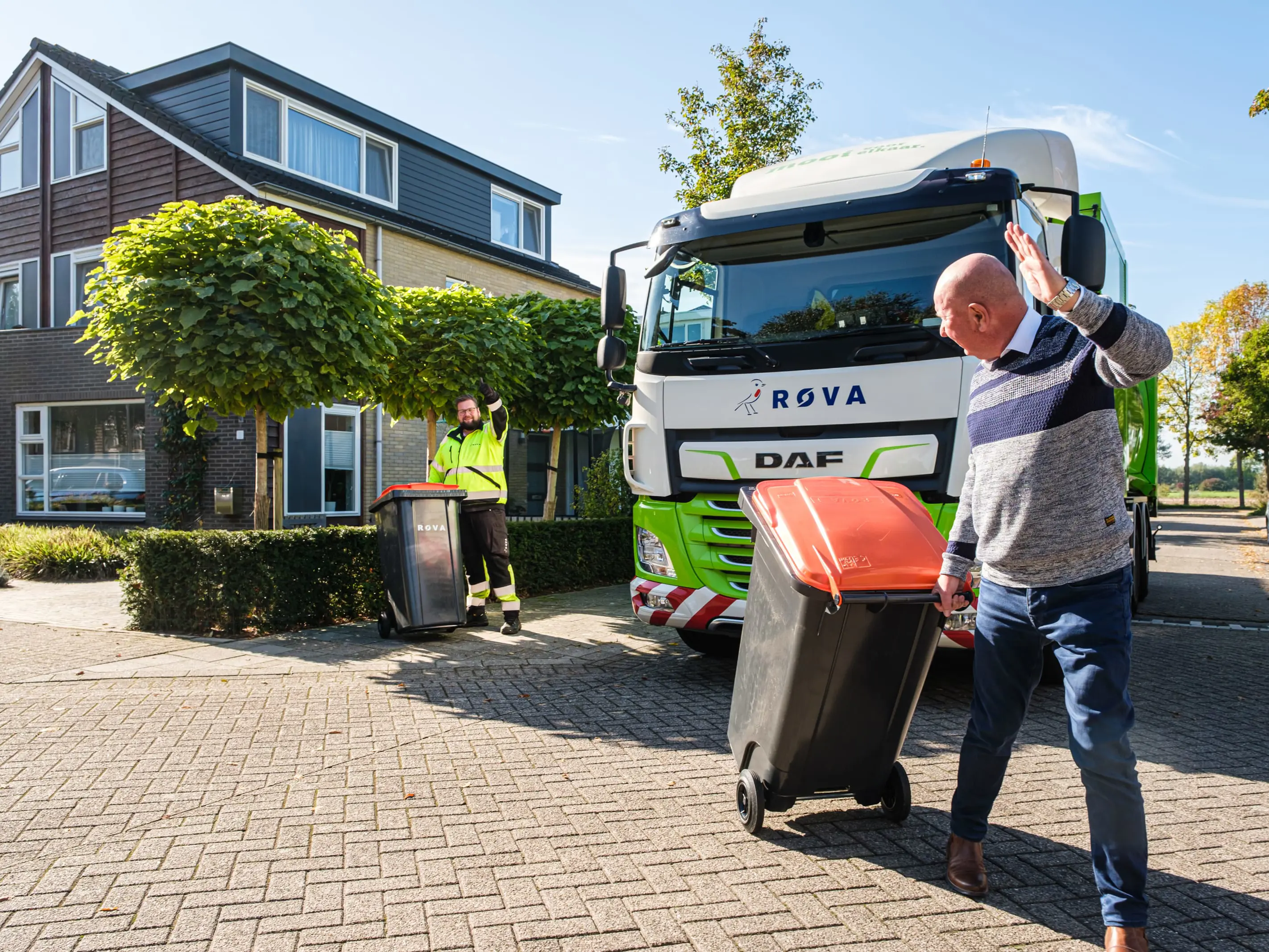 Sfeerfoto van een ROVA medewerker naast een ROVA afvalwagen, zwaaiend naar een inwoner met een groencontainer